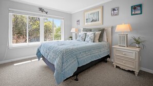 A bedroom with a large window, a bed with a blue bedspread, a nightstand with a lamp, and wall art. - Memories are Made Here (Hilton Head Island)