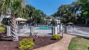 A fenced pool area with a clear blue pool, surrounded by lush greenery and a paved walkway. - Memories are Made Here (Hilton Head Island)