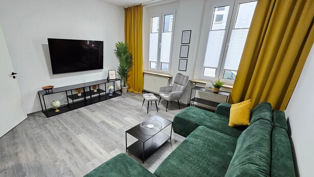 A modern living room with a large flat-screen TV, a green sofa, a coffee table, and a side table with decorative items. - Apartment 'Relax In Der Röntgenstadt' with Wi-Fi (Remscheid)