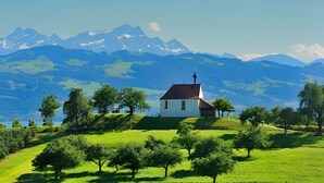 Un edificio blanco con techo rojo sobre una colina cubierta de hierba, rodeado de árboles y montañas al fondo.