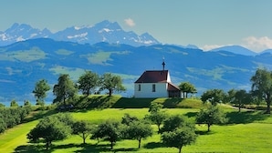A white building with a red roof on a grassy hill, surrounded by trees and mountains in the background.