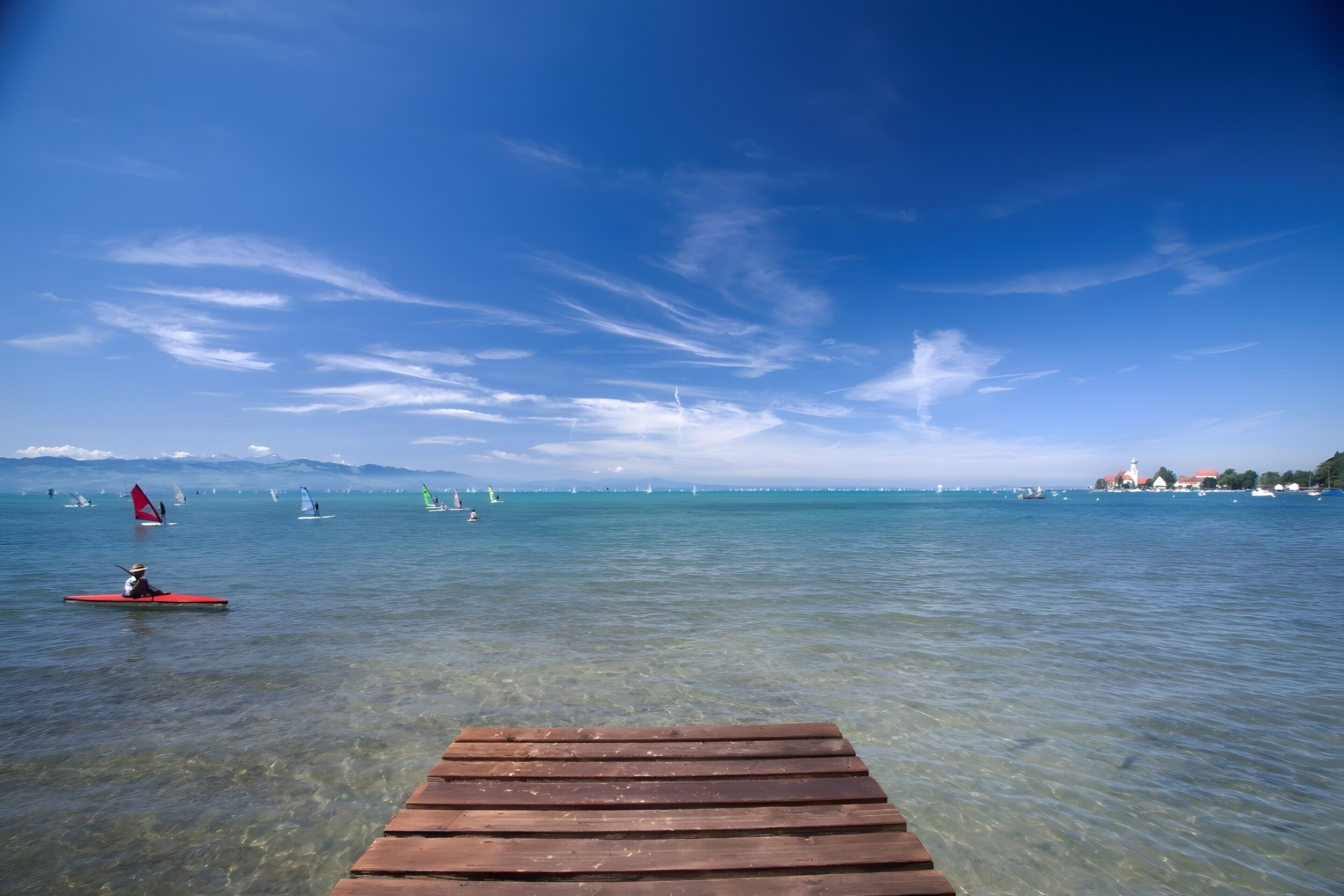 A wooden pier extending into a calm sea with sailboats and a distant shoreline.