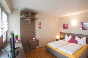 Quarto de hotel com uma cama, uma escrivaninha, uma televisão e uma pequena prateleira com objetos decorativos.