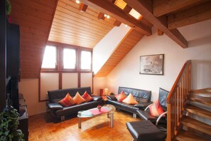 A loft-style living room with a wooden ceiling, black leather sofas, a glass coffee table, and a wooden staircase.