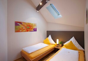Pequeno hotel com um quarto único, duas camas, ventoinha no teto, aparelho de ar condicionado e um quadro de flores na parede.