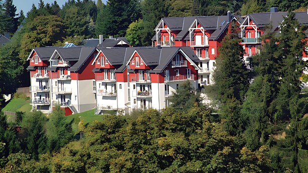 A cluster of red and white buildings with multiple balconies, surrounded by dense green trees.