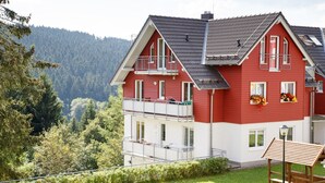 A red and white two-story house with a balcony, surrounded by greenery and a forested hillside.