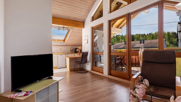 Una sala de estar moderna con una televisión de pantalla plana, suelo de madera, una zona de comedor con mesa y sillas, y ventanas amplias con vistas al exterior.