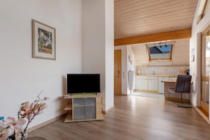 Un interior moderno con un televisor de pantalla plana sobre un soporte de madera, una cocina con armarios blancos y una silla de comedor.