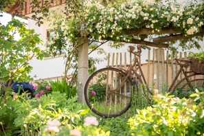 Een fiets leunt tegen een houten hek in een tuin met bloeiende planten en een wit gebouw op de achtergrond.