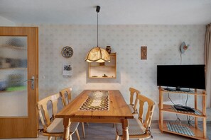 Comedor con mesa de madera, sillas, lámpara colgante, televisor sobre un mueble de madera y papel tapiz estampado.