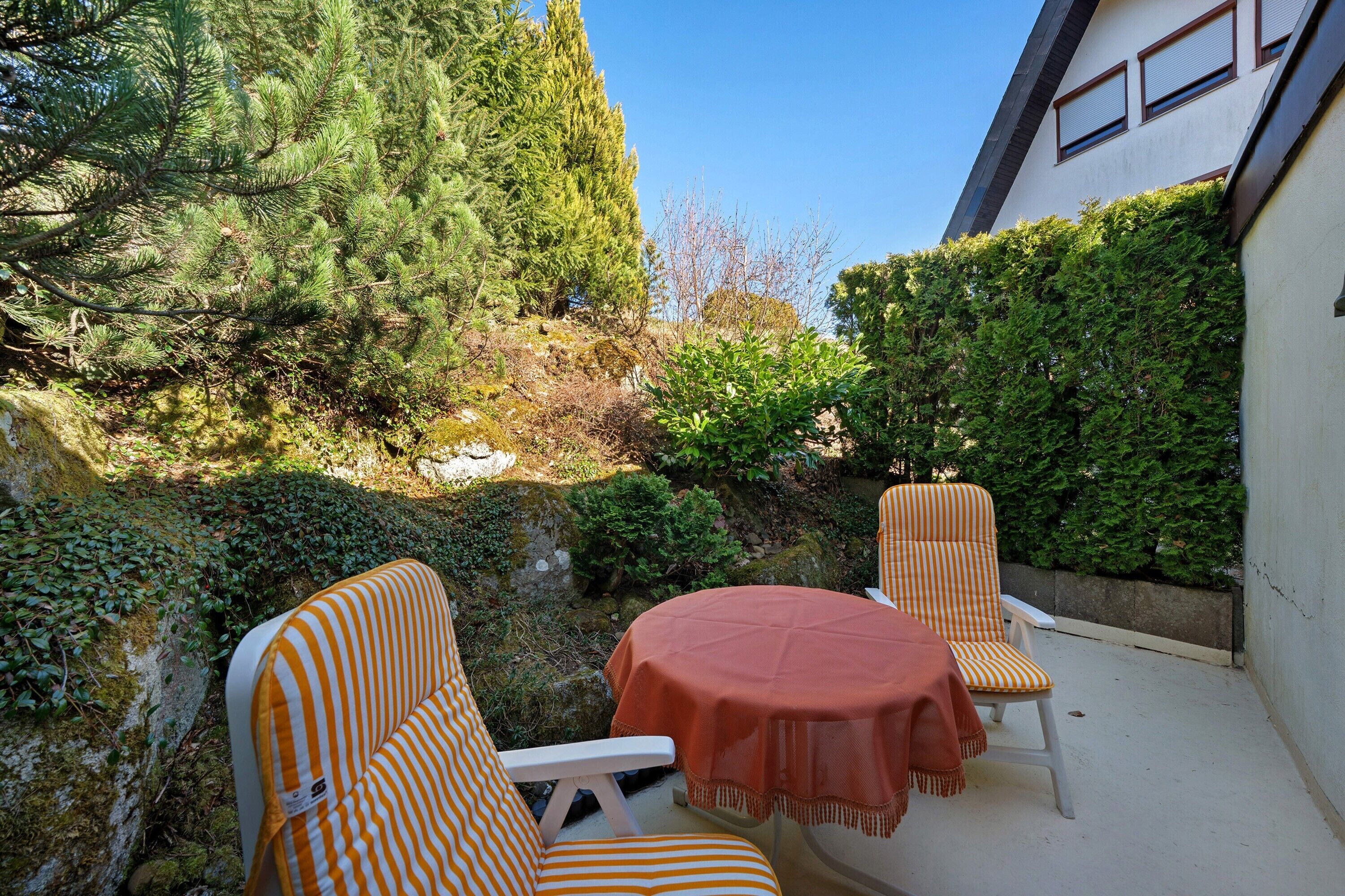 An outdoor seating area with two striped chairs and a table, surrounded by greenery and a building.