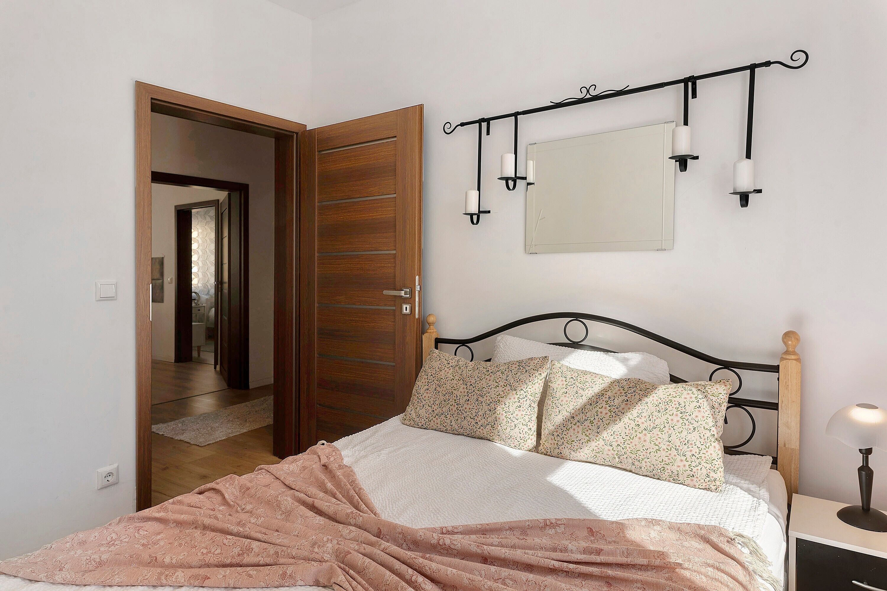 A bedroom with a wooden door, a bed with floral bedding, a nightstand with a lamp, and a wall-mounted light fixture.