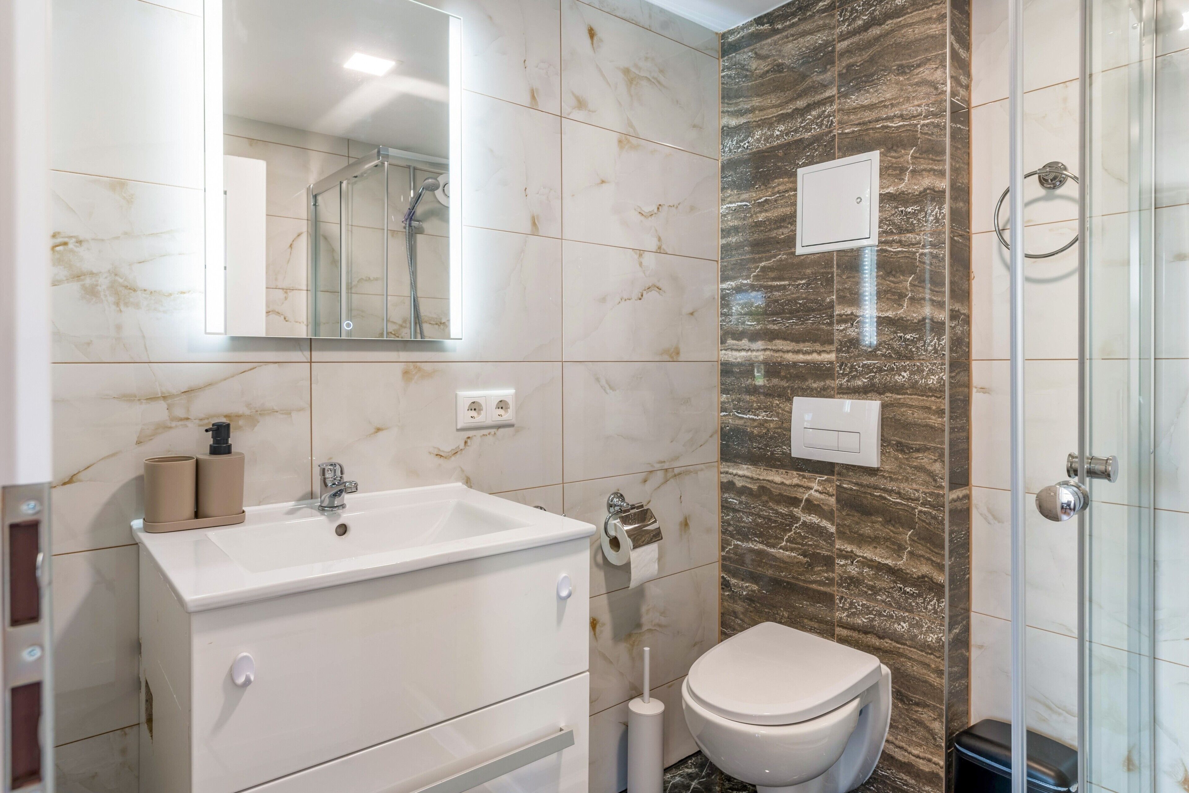 Un baño moderno con lavamanos blanco, espejo y retrete, paredes de mármol y cabina de ducha de vidrio.