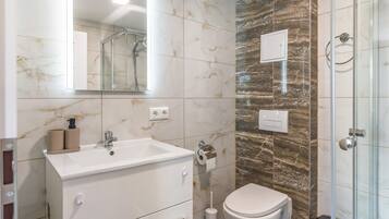 Un baño moderno con lavamanos blanco, espejo y retrete, paredes de mármol y cabina de ducha de vidrio.