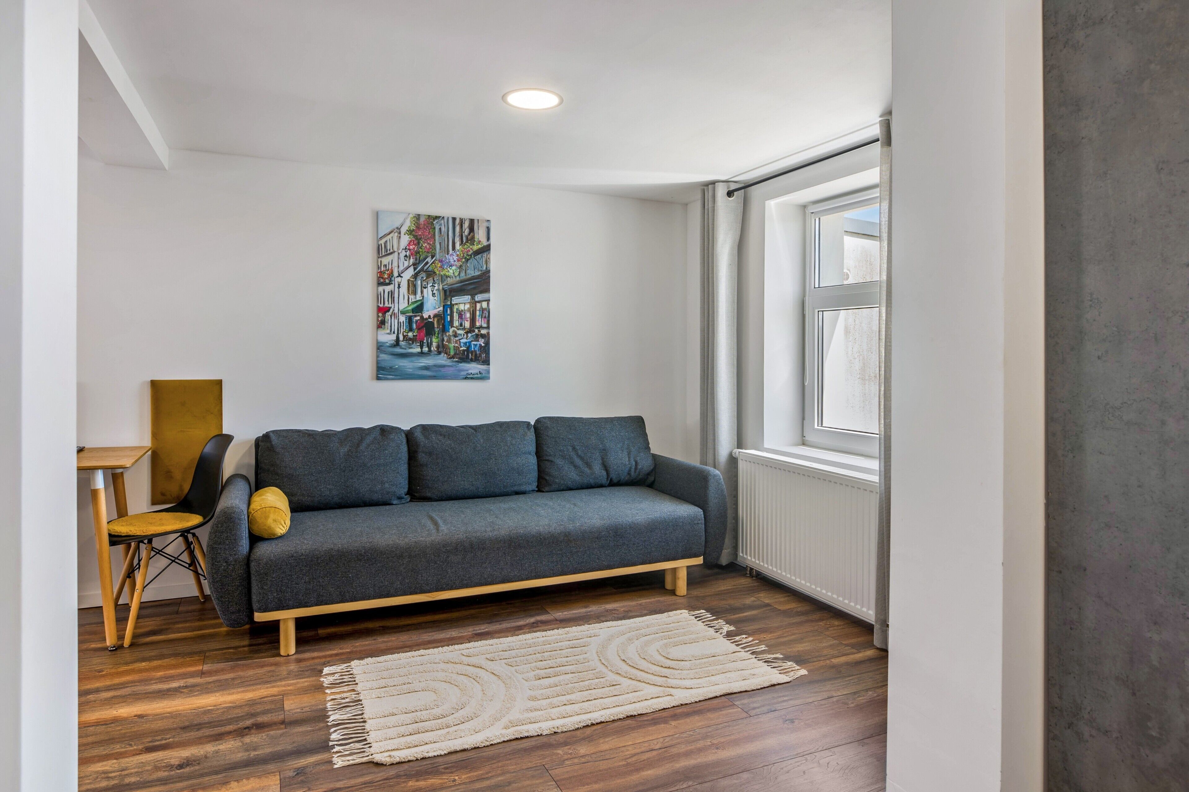 Un salon moderne avec un canapé gris, une table basse en bois, un fauteuil jaune, un tapis blanc et un tableau représentant un paysage urbain accroché au mur.