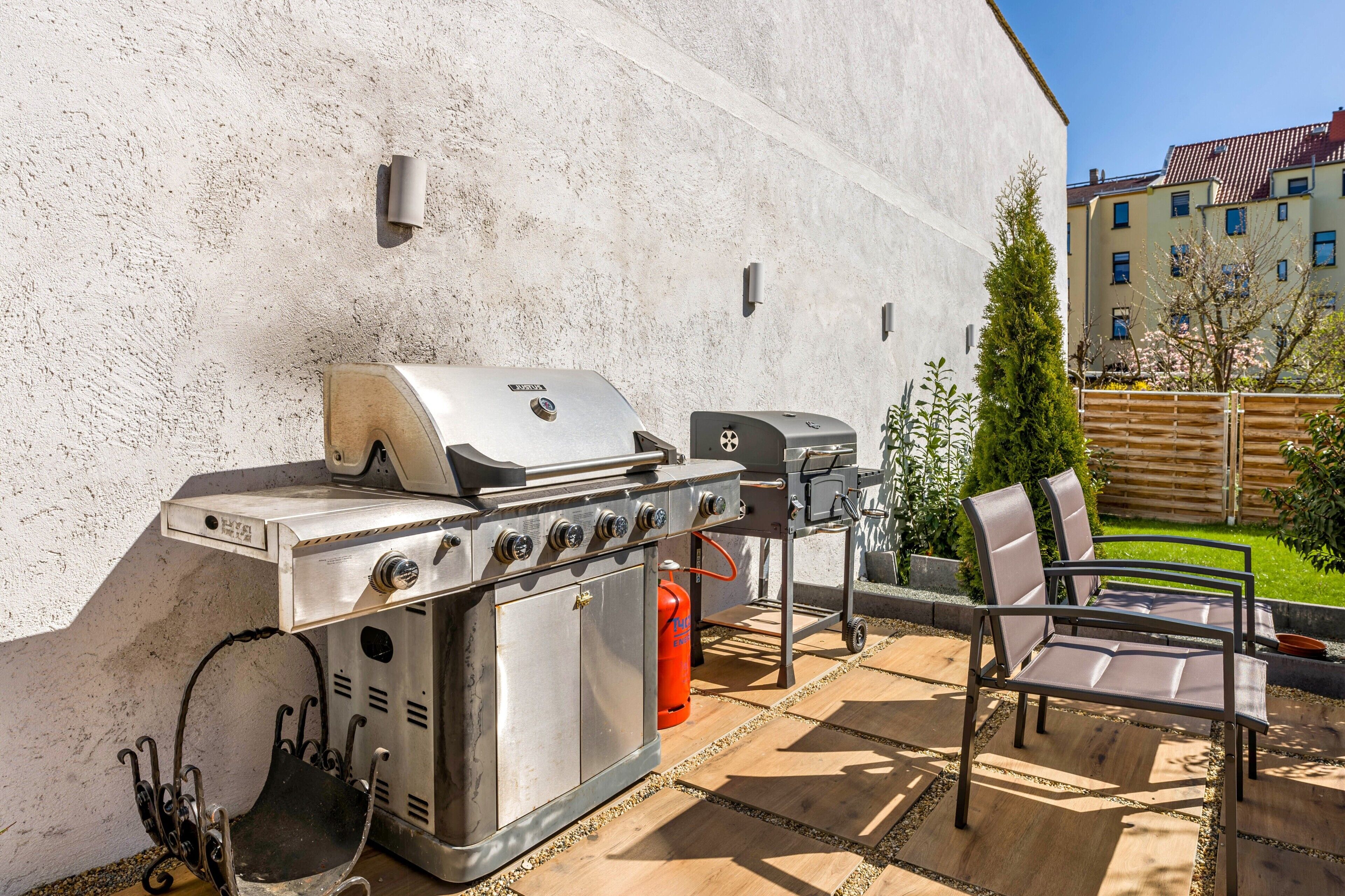 En udendørs terrasse med grill i rustfrit stål, en gasdåse og et siddeområde med stole.