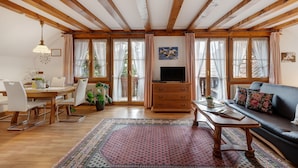 A spacious living area with wooden beams, a dining table, chairs, a sofa, a TV, and large windows.