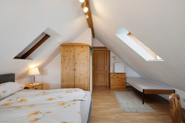 Une chambre avec un plafond en pente, un lit, une armoire en bois, une table de chevet avec une lampe, un banc et une petite fenêtre.