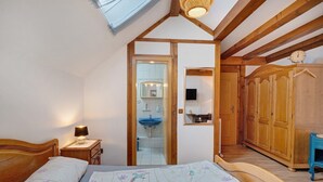 A bedroom with a skylight, wooden ceiling beams, a bed with floral bedding, a bedside table with a lamp, a bathroom with a sink and mirror, and a wardrobe.