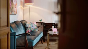 A room with a sofa, a standing lamp, a small table with a bottle and a card, and a painting on the wall.