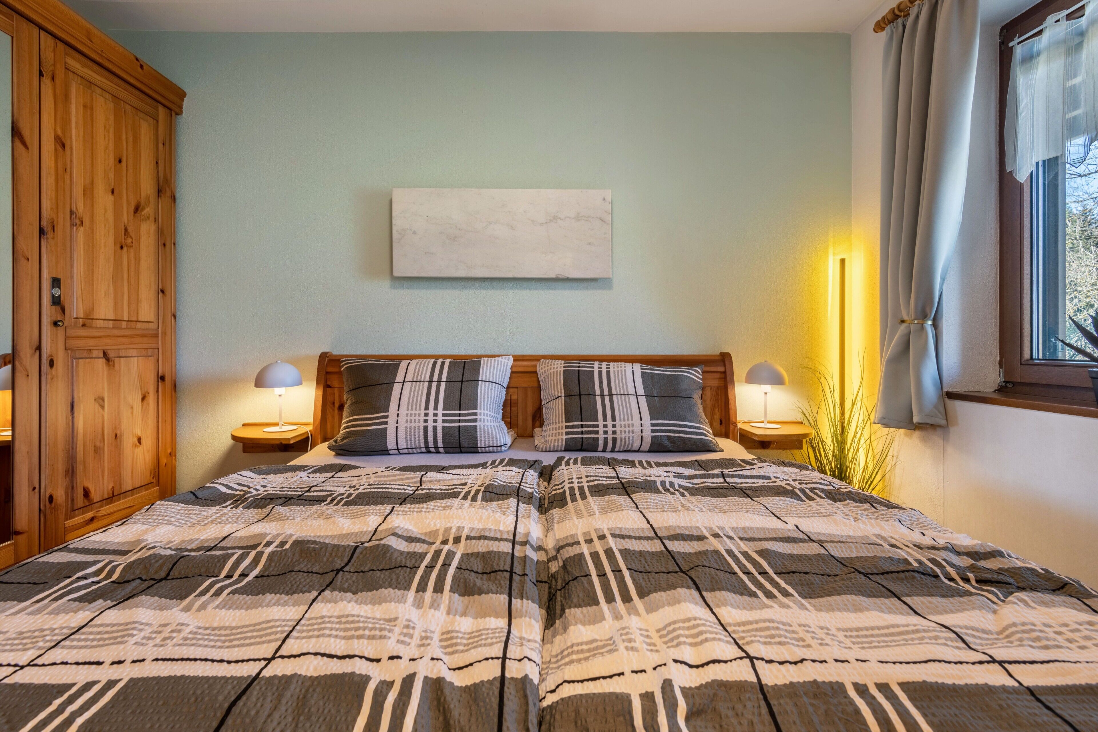 Een slaapkamer met een bed, twee nachtkastjes, een houten garderobe, een schilderij aan de muur en een raam met gordijnen.