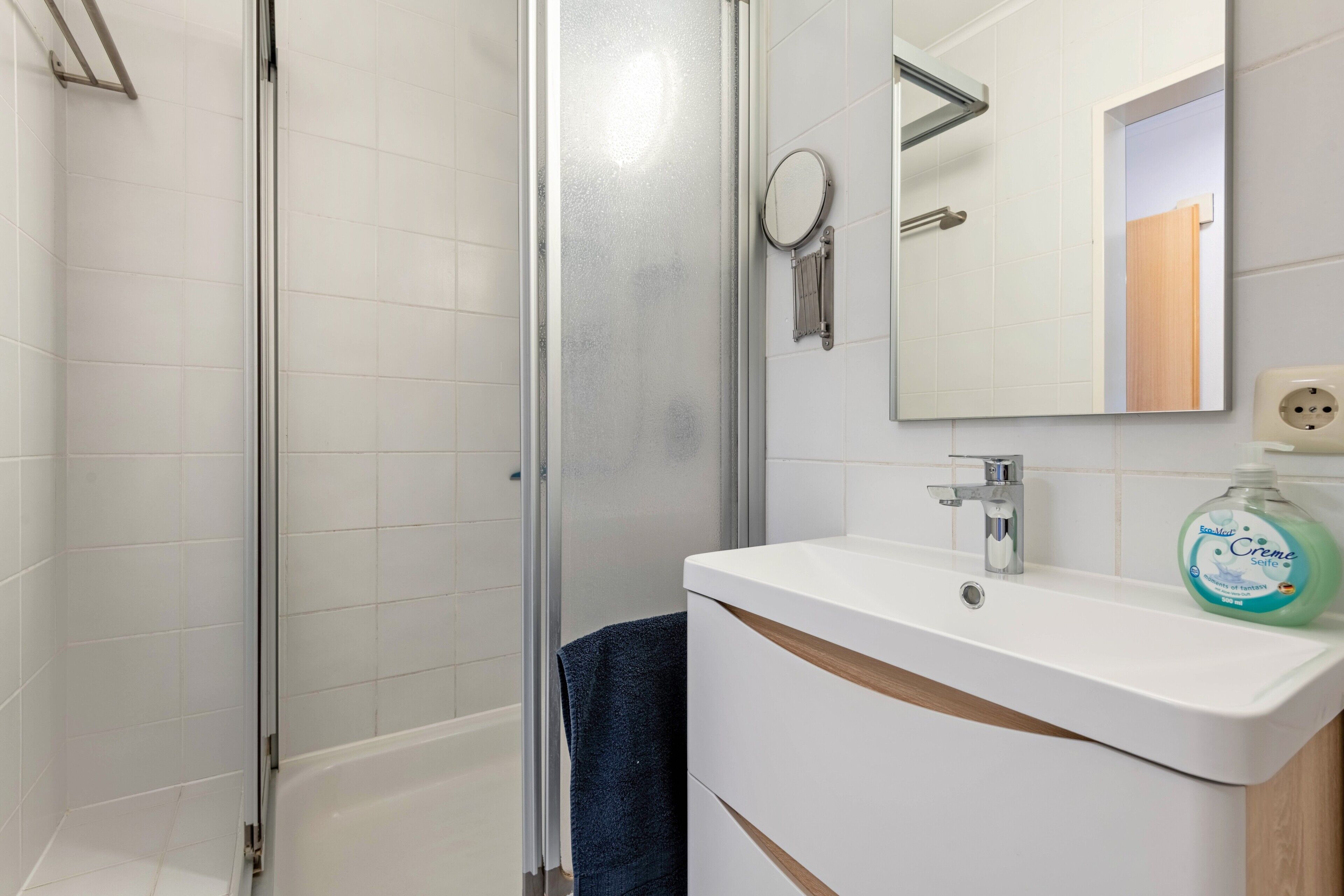 A bathroom with a glass shower enclosure, a white sink with a mirror, and a bottle of hand soap.