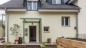 Ein zweistöckiges Haus mit grüner Tür, kleinem Fenster und Gartenanlage mit Pflanzen und einer Holzbank.