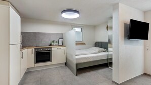 Ein gemütliches Zimmer mit Küchenzeile, einem Bett, einem Flachbildfernseher an der Wand und einem Fenster mit Jalousien.