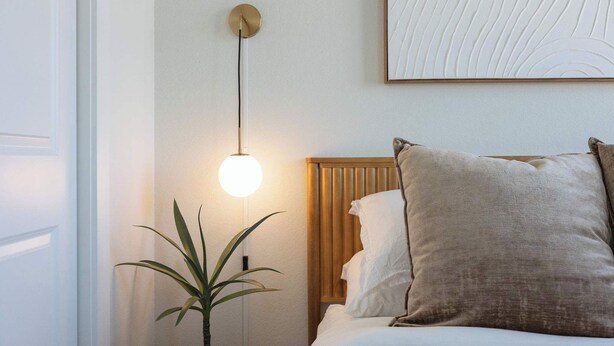 Quarto com cama, luminária de cabeceira, obra de arte na parede e planta em vaso.
