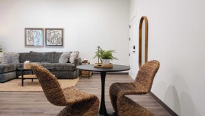 Uma sala de estar moderna com um sofá cinzento, duas cadeiras de vime, uma mesa pequena com uma planta e arte abstrata na parede.