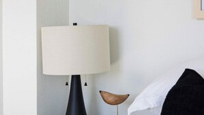 Ein ordentlich bezogenes Bett mit weißen Leinen, ein schwarzer Nachttisch mit Lampe und ein kleiner Holztisch daneben.