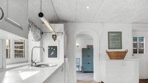 A modern kitchen with white cabinetry, a stainless steel sink, and a hanging light fixture. There is a blue chair and a basket on a white cabinet.