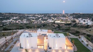 Un edificio moderno con varias estructuras blancas, una zona pavimentada con palmeras y una vista distante de un pueblo con una torre destacada.