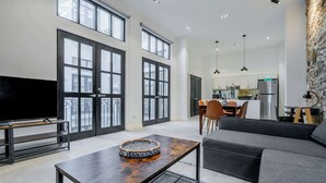Amplia sala de estar con un televisor grande, una mesa de centro de madera y un sofá gris. Se ven ventanas grandes y una cocina al fondo.