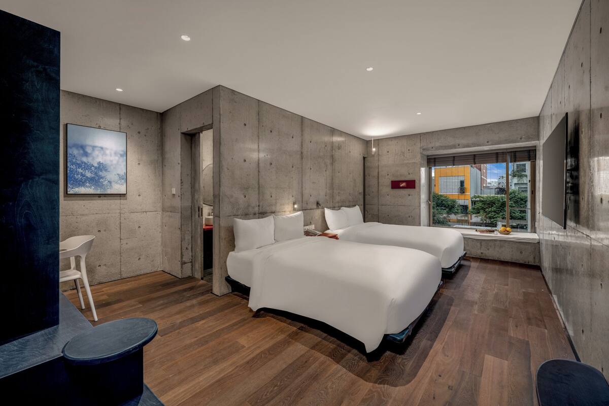 A modern hotel room with two beds, a small desk, and a view of the city.
