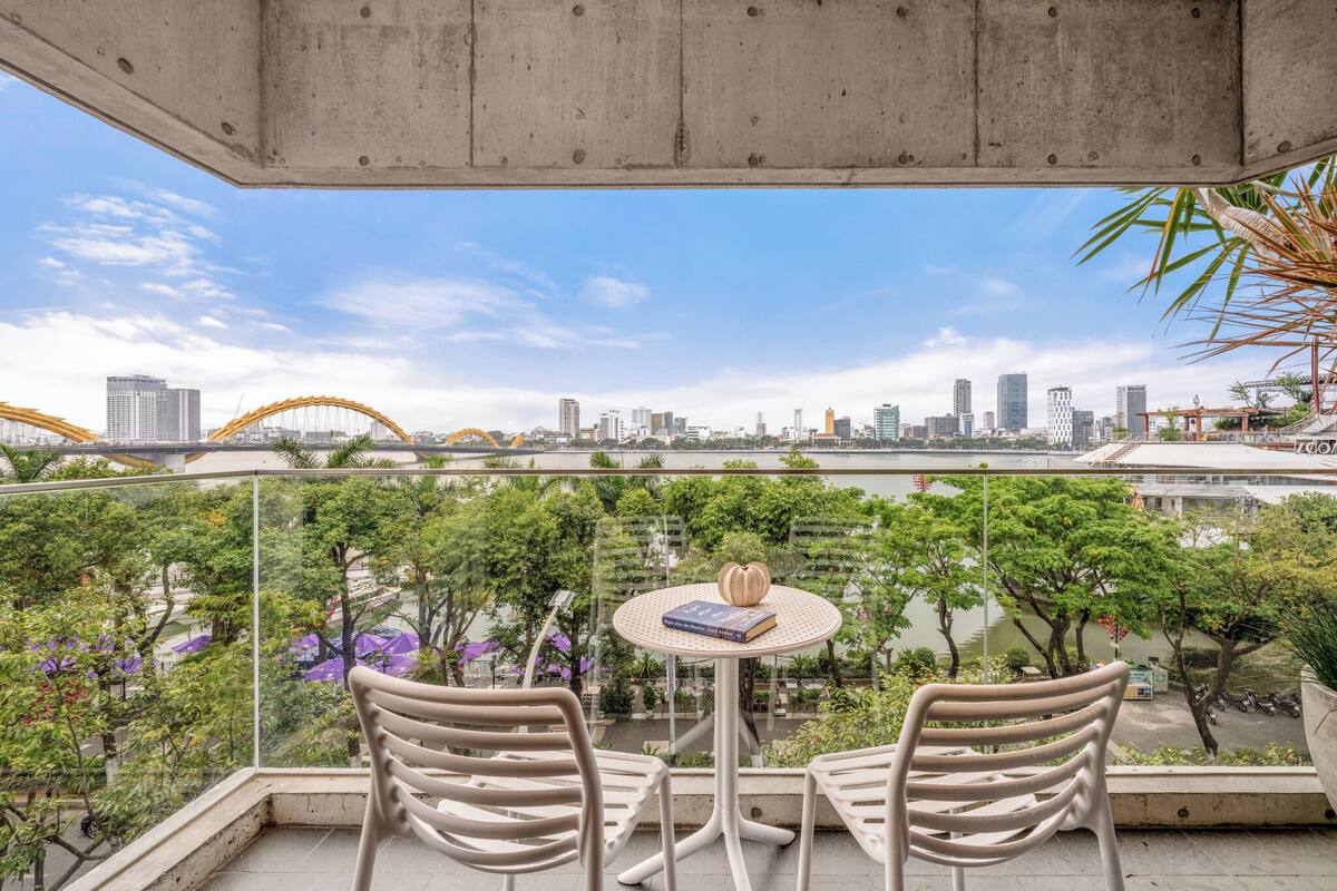 A balcony with two chairs, a small table with a book and a decorative item, and a view of a cityscape with buildings and a bridge.