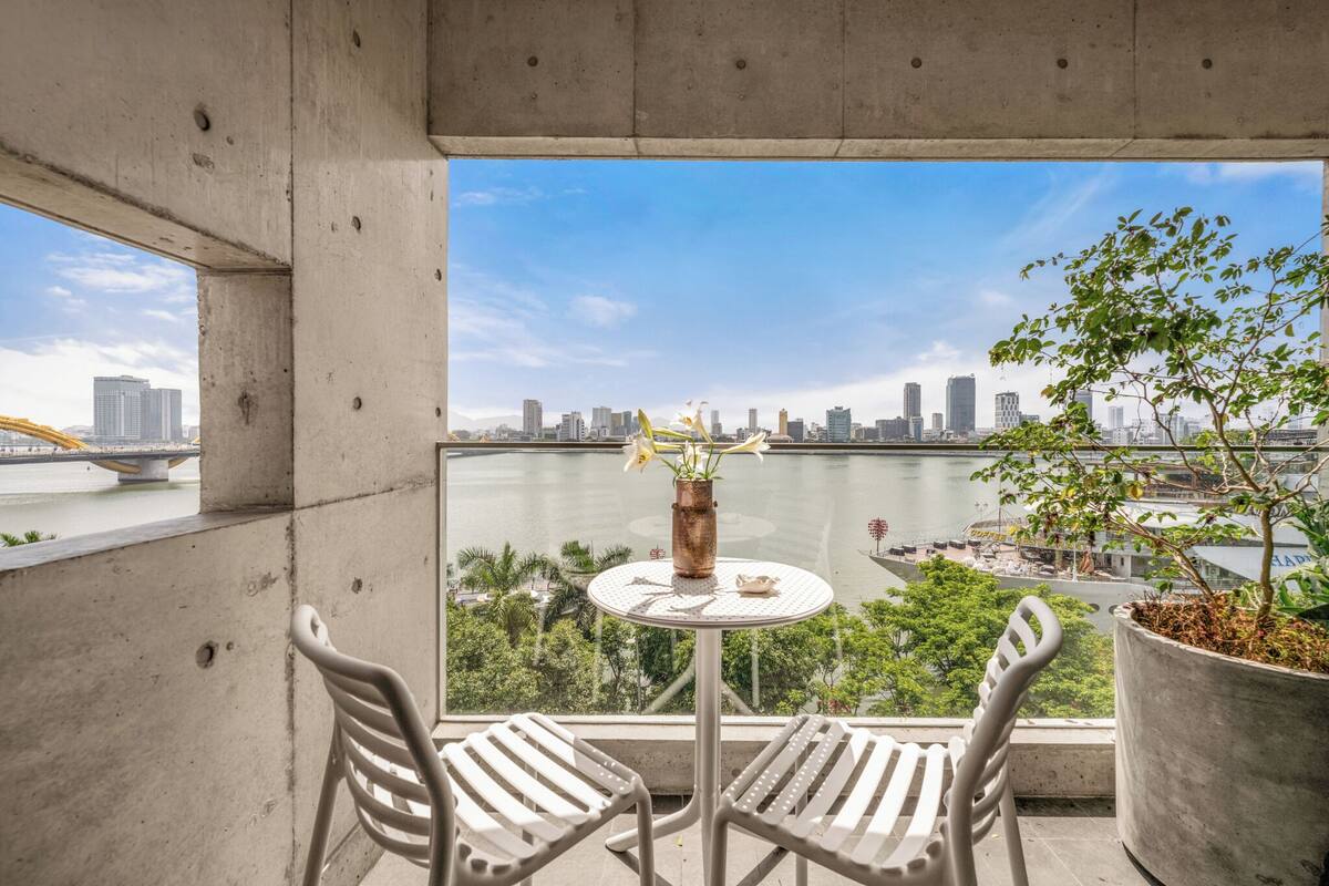 A balcony with two chairs and a small table, overlooking a river and a city skyline.