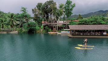 A river with a person paddleboarding, surrounded by lush greenery and a building with a thatched roof.