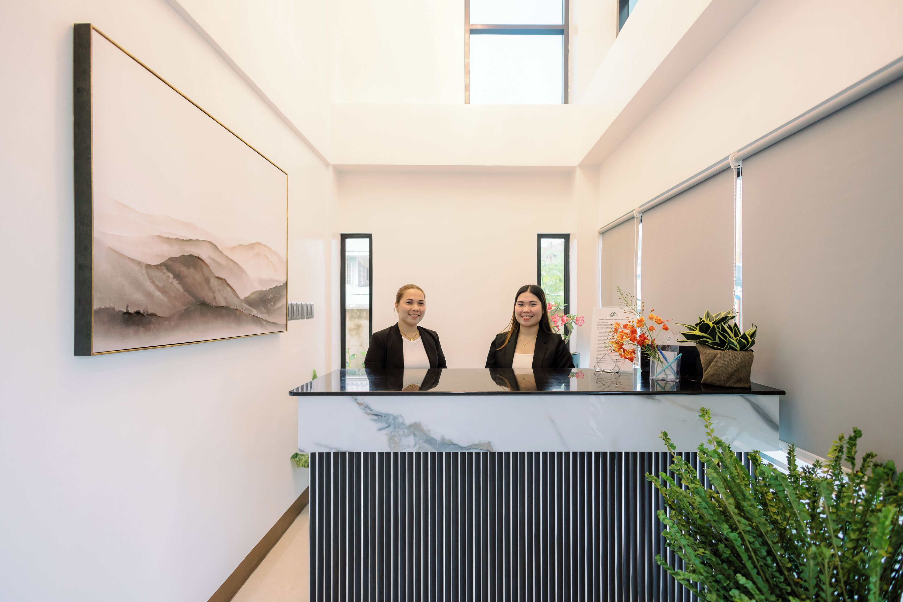 A reception desk with two staff members, a large abstract painting, and indoor plants.
