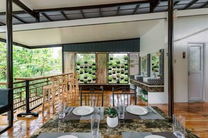 Uma área de refeições com uma mesa posta para dois, rodeada por cadeiras de madeira, vista para vegetação exuberante através de grandes janelas, e uma cozinha com painel de azulejos e bancada.