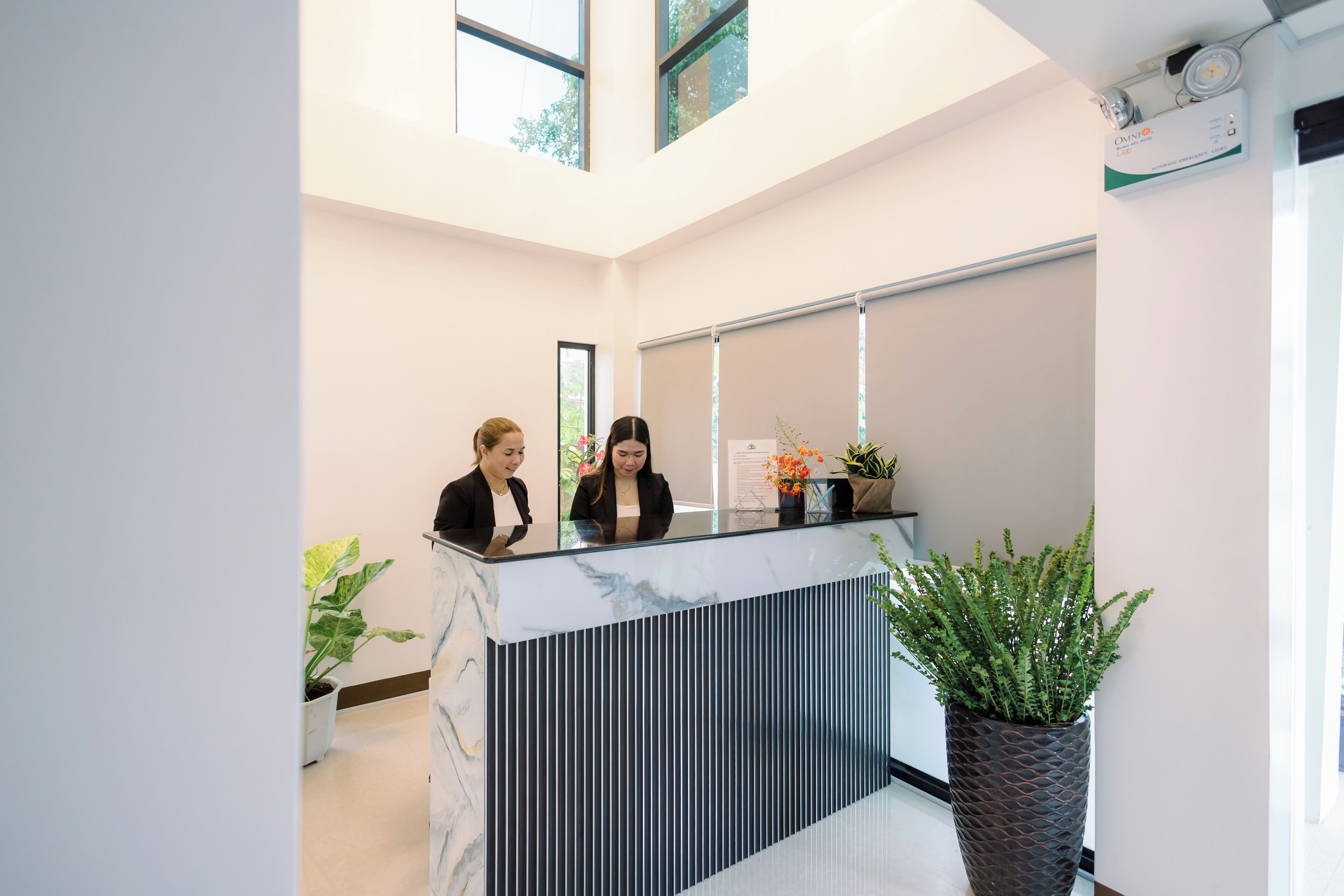Un espace d’accueil doté d’un comptoir en marbre, de deux réceptionnistes, de plantes en pot et d’une grande fenêtre.