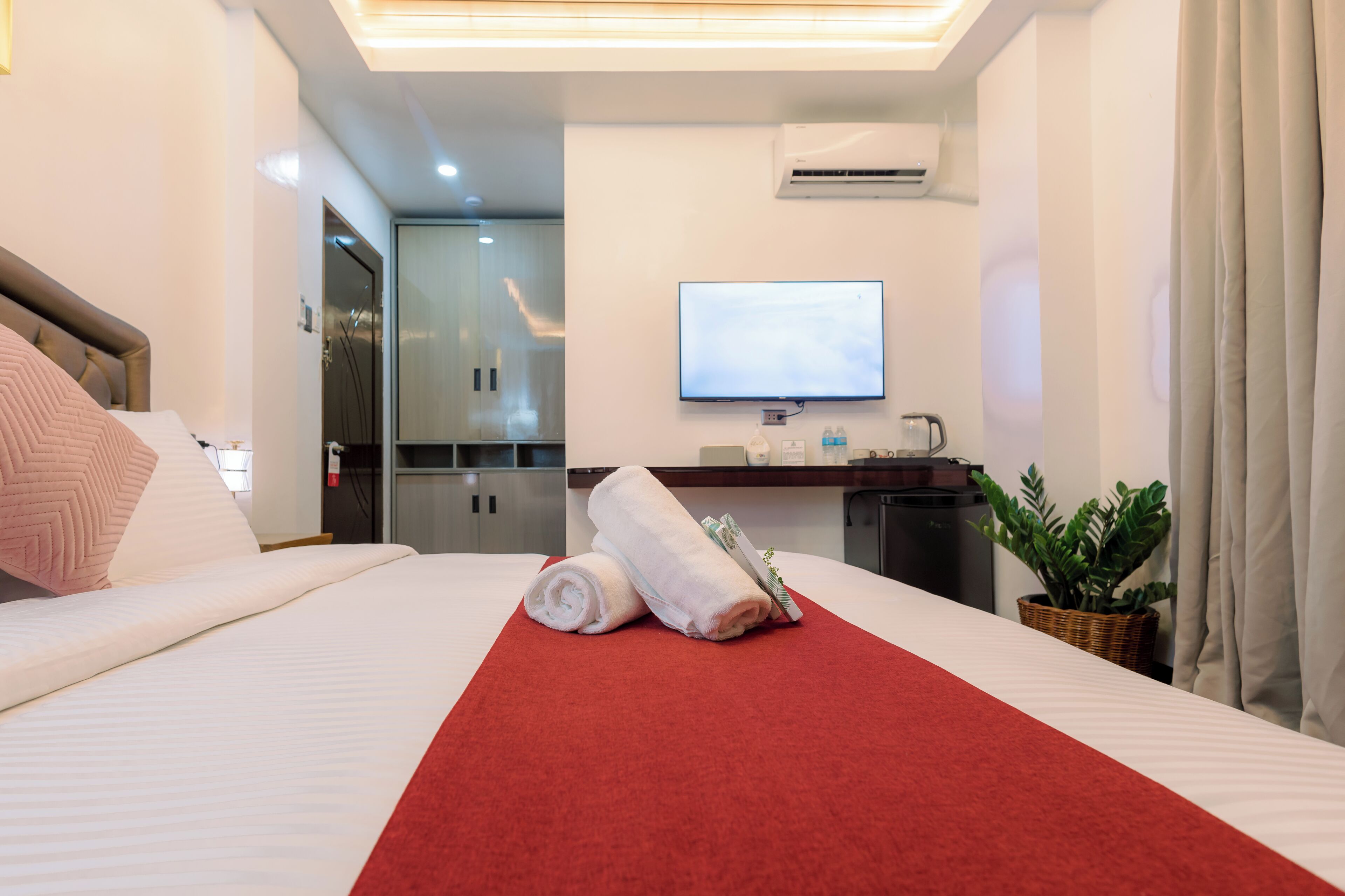 A hotel room with a neatly made bed, a red towel runner, a television mounted on the wall, and a small plant on a side table.