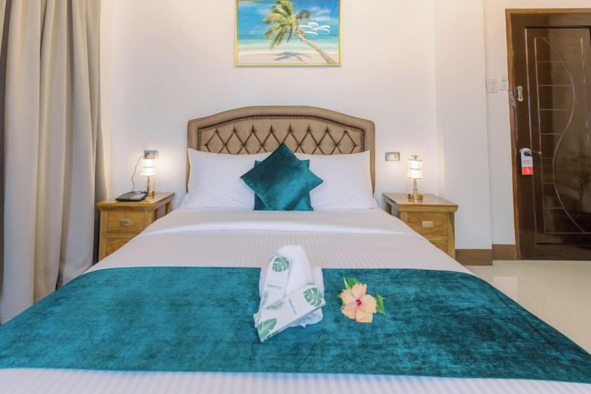 A neatly made bed with a teal bedspread, white pillows, and a single teal pillow. There are two bedside lamps on wooden nightstands, a framed picture of a beach scene on the wall, and a door with a glass panel.
