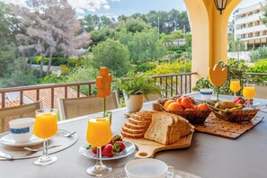 Un petit-déjeuner composé de jus d’orange, de pain et de fruits, servi sur un balcon offrant une vue sur la verdure et les bâtiments.