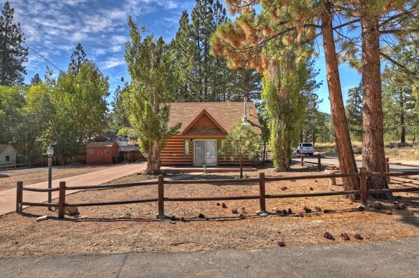 A small log cabin with a wooden deck and a fenced area, surrounded by tall pine trees. - Brother Bear Cabin (Big Bear City)