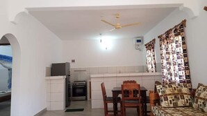 A compact kitchen with a refrigerator, stove, and microwave. There is a dining area with a wooden table and chairs. A ceiling fan is visible above the kitchen. - Kings Holiday resort watamu (Watamu)