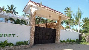 A gated entrance with a tiled roof, a stone wall with greenery, and palm trees in the background. - Kings Holiday resort watamu (Watamu)