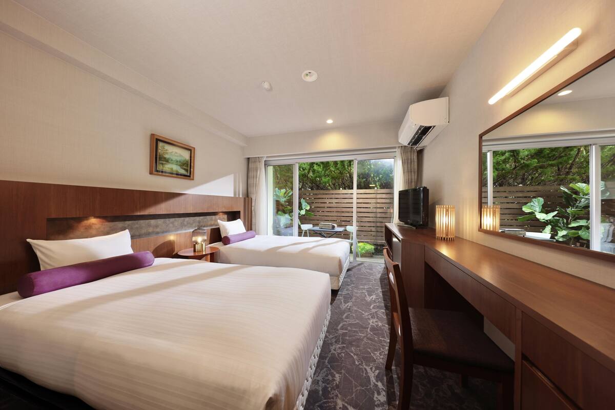 Une chambre d’hôtel avec deux lits, un bureau, une télévision et une grande fenêtre donnant sur un paysage verdoyant.