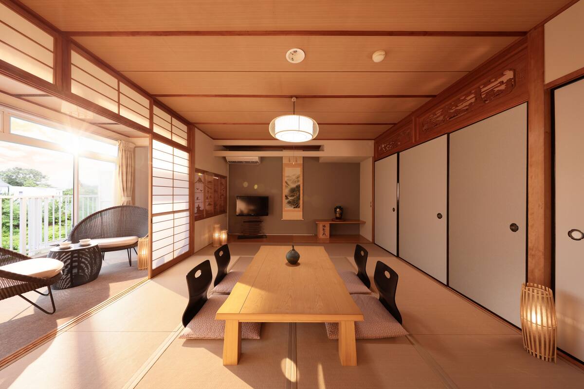 Une pièce de style japonais traditionnel, avec un sol recouvert de tatamis, une table à manger en bois, des chaises noires et un téléviseur en arrière-plan.